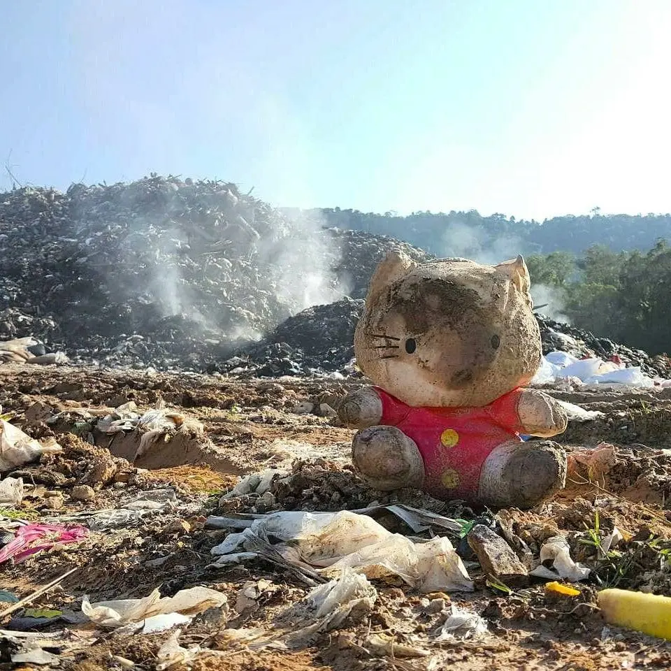 A charred Hello Kitty toy sits before a smoking mountain of trash